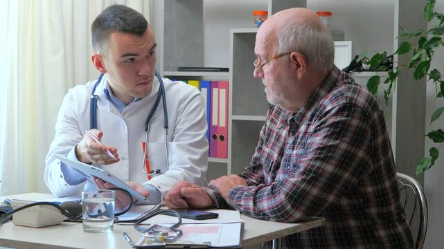 A young doctor in the office, receives an elderly man, explains his health condition, makes a diagnosis, prescribes appropriate treatment, and provides moral support to the elderly patient. Slow motio