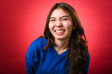 Fototapeta premium An Asian woman in a blue sweater smiles with a scrunched-up face, showing her teeth in a playful or cheeky expression. The background is bright red, and her long hair falls over her shoulders.