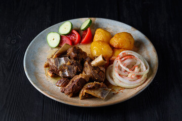fried lamb ribs, meat with baked potatoes in oil with fresh tomatoes, cucumbers and onions on a black wooden background