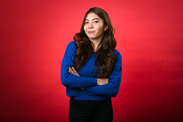 Asian woman with long, wavy brown hair is smiling while standing confidently with her arms crossed. She is dressed in a blue sweater and black pants, posing in front of a red background