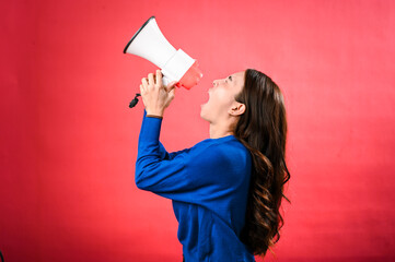 An Asian woman wearing a blue sweater is holding a red and white megaphone while speaking into it. She appears to be shouting or making an announcement. The background is solid red