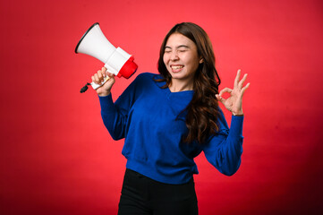 An Asian woman wearing a blue sweater is holding a red and white megaphone while speaking into it. She appears to be shouting or making an announcement. The background is solid red