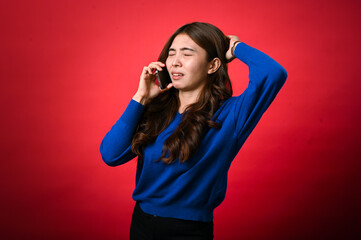 An Asian woman in a blue sweater holds a phone to her ear, with her eyes closed and an expression of frustration or sadness. She stands against a red background, showing a disappointed mood