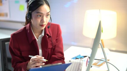 Young asian woman wearing a headset and a red jacket is working at her desk. She is having a video call and seems to enjoy it