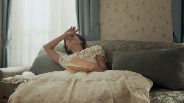 Medium shot of shocked 19th century young woman fainting on upholstered sofa after getting bad news by letter written on parchment paper