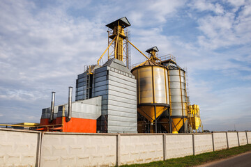 Obraz premium large grain elevator with multiple cylindrical metal silos, structural elements, and a complex system of pipes. It stands against a blue sky, with a field in the foreground after harvest