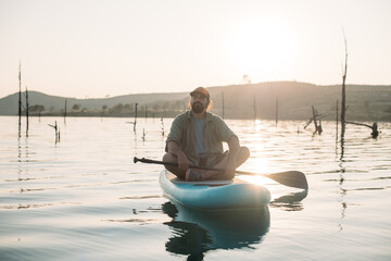 A man is surfing on a sap board on calm water at sunset.