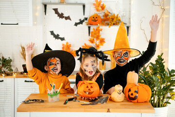 Halloween, kids boys and a girl in festive costumes and makeup are preparing for Halloween at home in the kitchen by coloring and carving pumpkins and scaring everyone