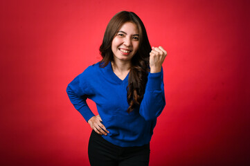 An Asian woman in a blue sweater and black pants is standing against a red background, smiling confidently while raising her fist in a gesture of strength and victory, her other hand on her hip