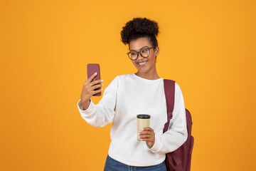 A cheerful young woman smiles as she takes a selfie with her phone. She holds a coffee cup in one...