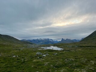 A serene, remote landscape of green valleys and rocky terrain, with snow-capped mountains reflecting in calm waters beneath an overcast sky, radiating peaceful solitude.