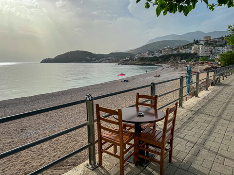 Small table and chairs are set up in a cafe on promenade overlooking sandy beach Potami in tourist town Himare, Vlore, Albania. Sunset at Ionic Mediterranean sea. Scenic vistas of Ceraunian mountains