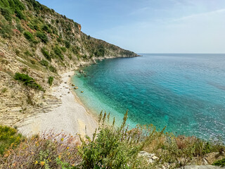 Selective focus on flowers with scenic view of secluded pebble beach Filikuri surrounded by dramatic cliffs in Himare, Albania. Crystal-clear turquoise water of Ionian Mediterranean sea. Tranquility