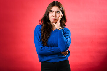 An Asian woman with long brown hair, wearing a blue sweater, poses with a thoughtful expression. She rests her chin on her hand while standing against a red background.