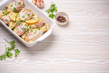 Raw uncooked chicken legs with seasonings, herbs, lemon in white ceramic casserole top view on light wooden rustic background. Preparing healthy meal with marinated chicken drumsticks. Space for text.