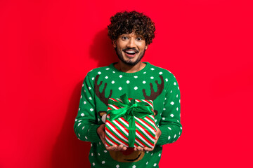 Portrait of nice young man hodl giftbox wear new year xmas sweater isolated on red color background