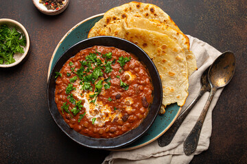 Traditional Indian Punjabi dish Dal makhani with lentils and beans in black bowl served with naan flat bread, fresh cilantro and two spoons on brown concrete rustic table top view.