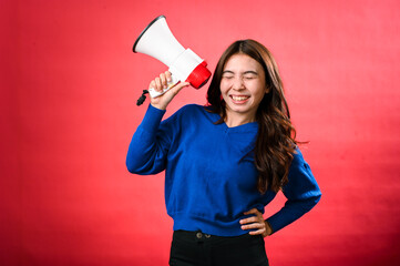 An Asian woman wearing a blue sweater is holding a red and white megaphone while speaking into it. She appears to be shouting or making an announcement. The background is solid red