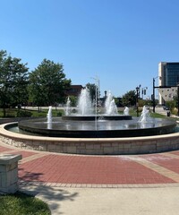 A large circular fountain in the middle of the city with jets of water shooting upwards, surrounded by brick paths and greenery, creating a serene public space.