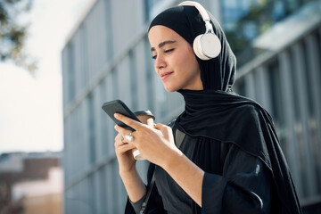 Smiling beautiful woman with hijab listening to music with headphones while using smartphone walking throught the city