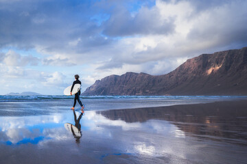 An athletic surfer walks the beach with a surfboard during sunset, famara beach, Lanzarote, Canary...