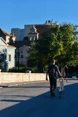 A bald, casually dressed man, with his back turned, pushing a bicycle down a street in a European city