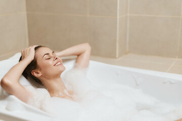 Relaxed young woman enjoying a bubble bath, smiling with her eyes closed, surrounded by foam in a...