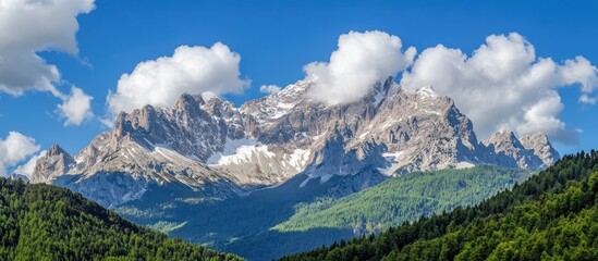 A majestic mountain range with snow-capped peaks and lush green forests in the foreground, under a clear blue sky with white clouds.