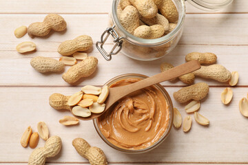Tasty peanut butter in bowl, nuts, jar and spoon on wooden table, flat lay