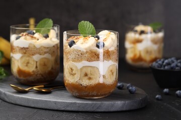 Tasty trifle dessert. Sponge cake, blueberries, banana and whipped cream in glasses on black table, closeup