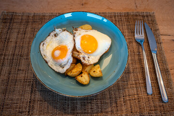 Lovingly prepared breakfast with potatoes and fried egg served on a blue plate at the wooden table.