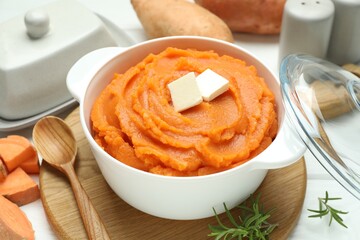Tasty mashed sweet potato with butter in bowl, spoon, fresh vegetables and rosemary on table, closeup