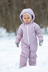 Side view toddler girl in light pink jumpsuit and a white fluffy hat walking in winter park. Children games in the snowy forest. Family winter vacation with a child. Vertical photo