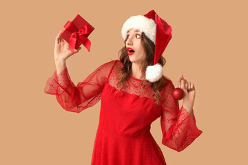 Surprised young woman in Santa hat with Christmas ball and gift box on brown background