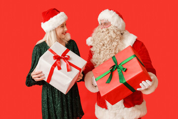 Santa Claus and beautiful mature woman with gift boxes on red background