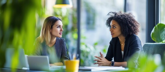 Two diverse young businesswomen sit at a table in a cafe, discussing ideas and collaborating on a project.