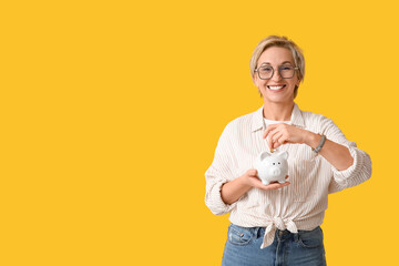 Beautiful mature woman putting coin into white piggy bank on yellow background