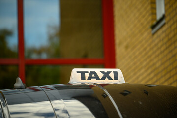 A noticeable taxi sign rests on the roof of a parked vehicle, reflecting the clear sky above. The background shows a brick building with large windows