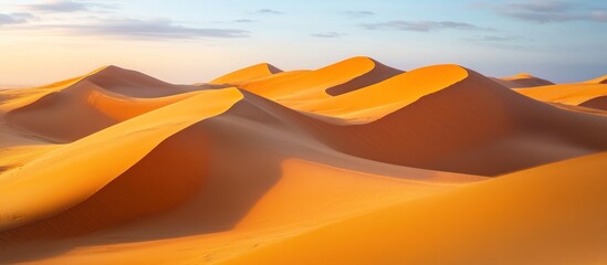 A panoramic view of golden sand dunes in the desert with a soft blue sky above.