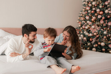 Tired parents with their son playing on a tablet on the bed. Two parents playing with their son in tablet at home. Part of a series