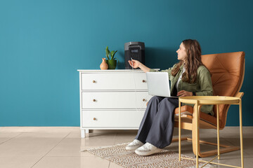 Young woman charging laptop with portable power station in armchair at home
