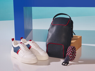 White sneakers, dark sports bag and knitted belt on decorative studio background for men. Red line and patterned design, black and white red belt, blue and white background.