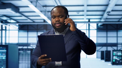 IT professional looking over data center checkup checklist paperwork, talking on phone in server farm. African american man surrounded by server rigs, chatting during telephone call, camera A