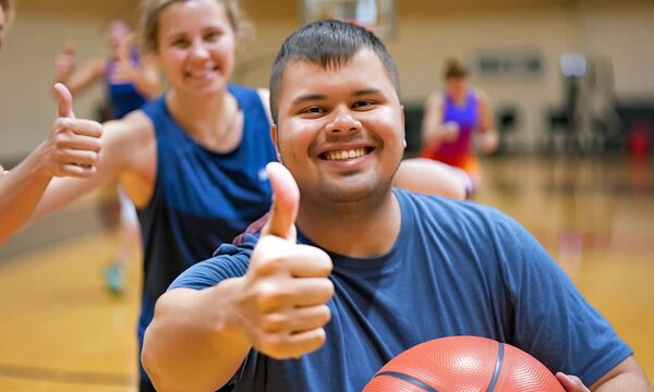 Young adult with down syndrome smiling and giving a thumbs up while holding a basketball in a lively gym environment