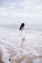 Beautiful graceful gentle girl with long dark hair in a white dress stands on the seashore, the waves wash her bare feet, her hair flutters in the wind