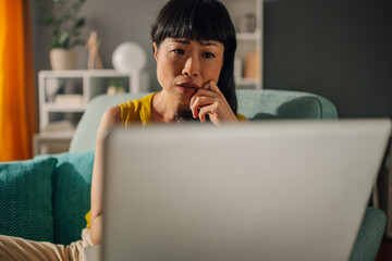 Asian woman thoughtfully looking at the laptop in her cozy living room