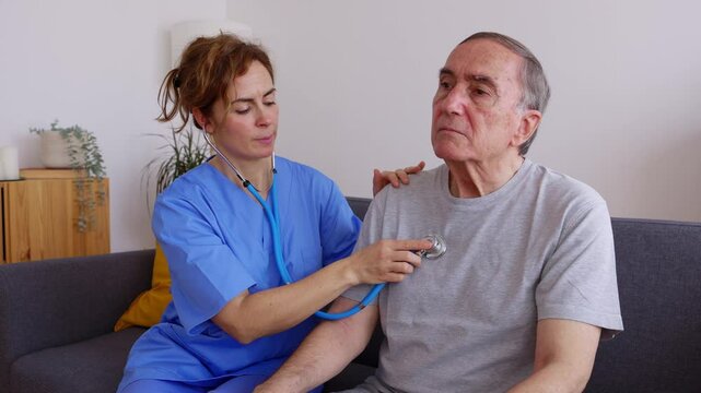 Young female doctor in blue uniform listening to disabled older patient heart sound with stethoscope during visit at home. Medical checkup and healthcare concept