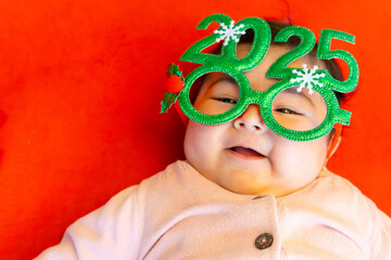 Greeting card Happy new year 2025, smiling Japanese baby celebrating new year with glasses numbers 2025 on red background with copy space. Funny postcard, natality and prosperity for next year.