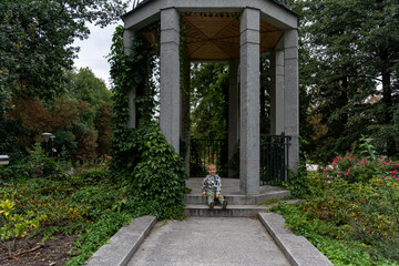Little boy sitting on steps of a stone gazebo covered in ivy in a peaceful park garden. Concept of childhood curiosity, nature exploration, and serene outdoor spaces. High quality photo