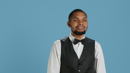 Friendly bellman porter with suit and tie waving at hotel guests, greeting people and offering excellent concierge services. Young employee saying hello to clients against blue background. Camera A.